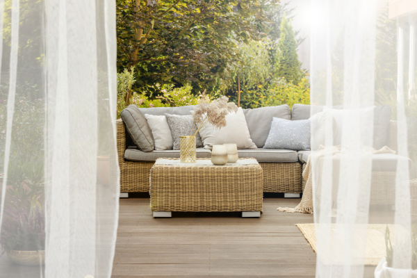 Elegant wicker patio furniture set with grey cushions on a wooden deck surrounded by garden greenery.